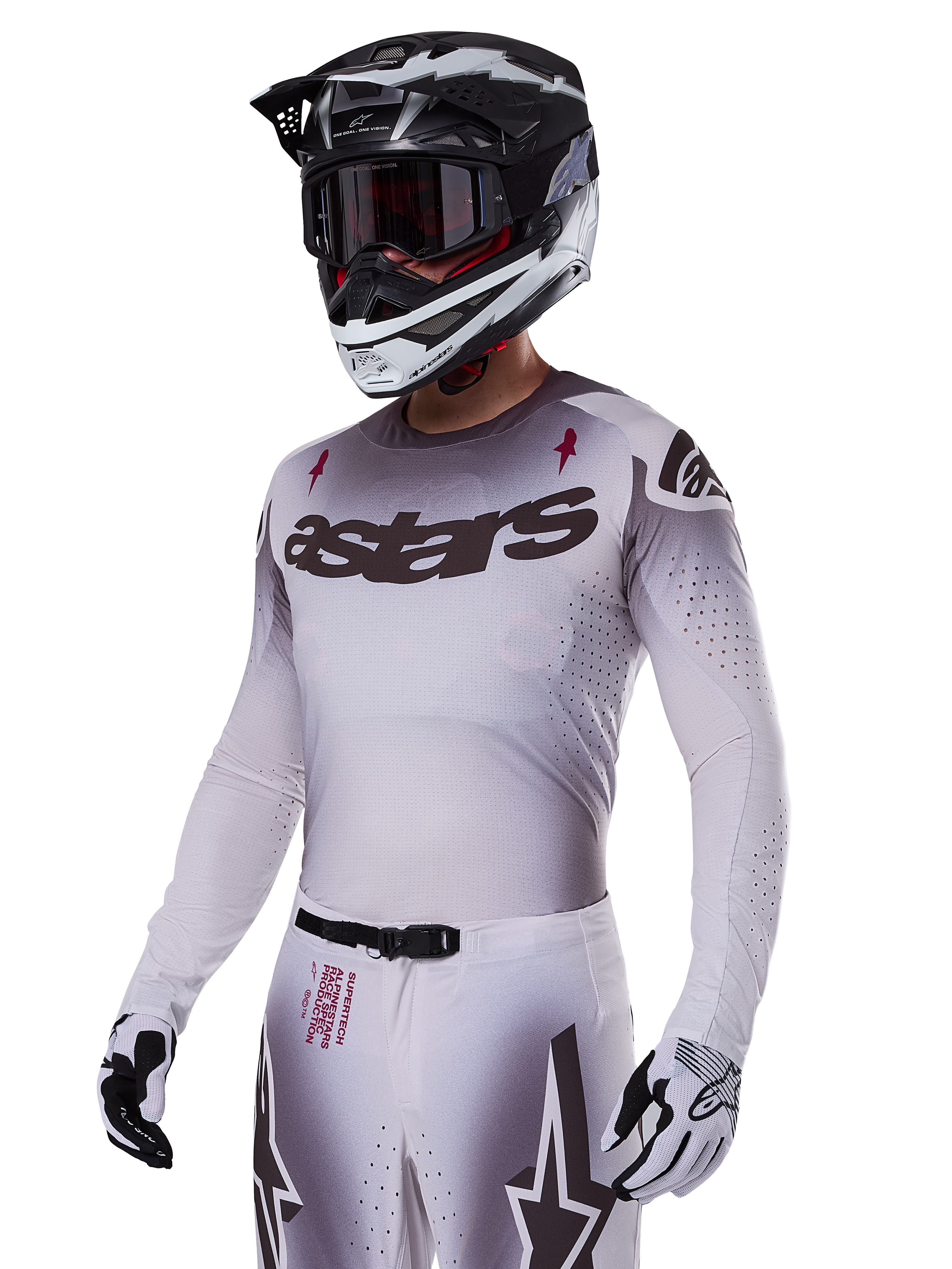 Motorcrosser die de Alpinestars 2025 Supertech Maker Gear Set in het wit draagt, met een jersey met grijs kleurverloop, het zwarte astars-logo en lasergesneden perforaties, bijbehorende broek met magnetische gesp en ster-graphics, gedragen met een zwart-witte helm en handschoenen voor een complete off-road race-look.
