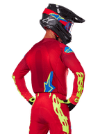 Achteraanzicht van een motocrossrijder die de Alpinestars 2025 Supertech Maker Gear Set in het rood draagt, bestaande uit een rode jersey met neongele branding op de mouwen, een bijpassende rode broek, een zwarte helm met blauwe en rode accenten en bijpassende zwart-gele handschoenen.