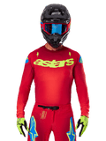 Alpinestars 2025 Supertech Maker Jersey in Bright Red Yellow Fluo, met geel 'astars' logo op de borst en blauwe sterrendesigns op de schouders, gedragen met bijpassende broek, zwarte helm, blauwe bril en gele handschoenen, professionele motorcross racekleding