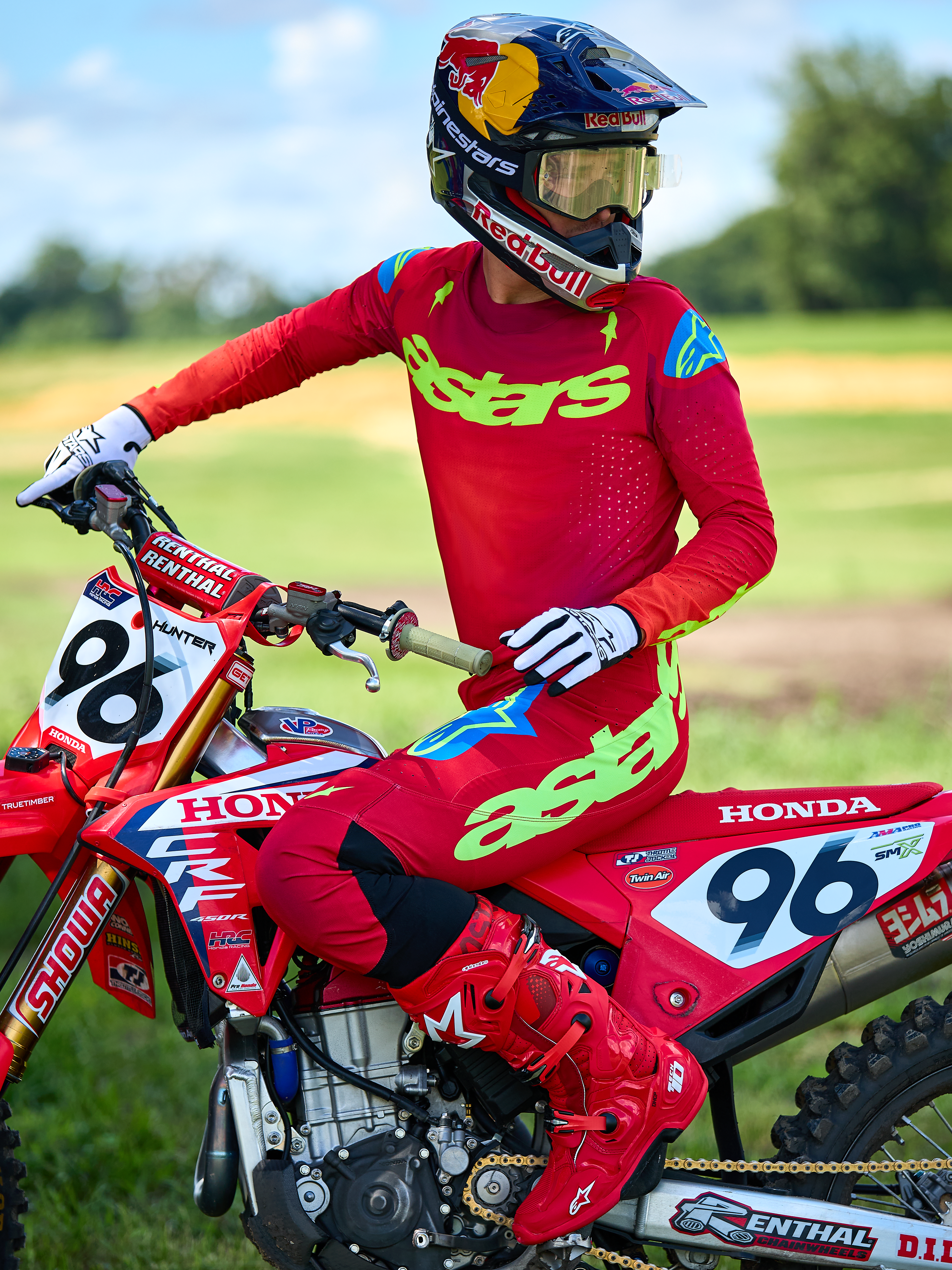 Motorcrosser op een rode Honda motorfiets die de Alpinestars 2025 Supertech Maker Gear Set in het rood draagt, voorzien van een rode jersey met neongroene 'astars' branding en blauwe schouderaccenten, bijpassende rode broek, effen rode laarzen, witte handschoenen en een Red Bull-helm, professionele race-uitrusting voor off-road prestaties