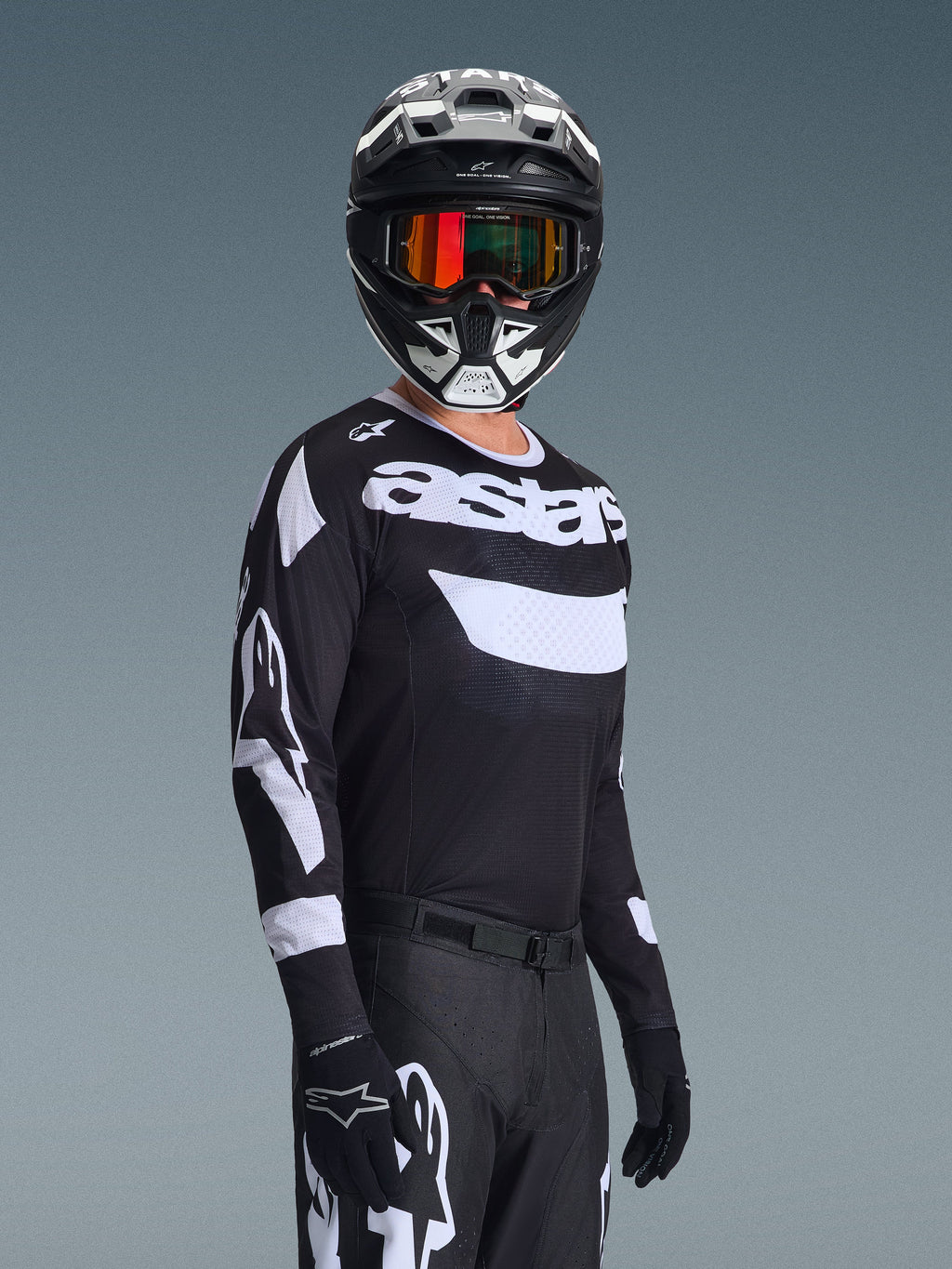 Motorcrosser in de Alpinestars 2026 Racer Air Riway Jersey, zwart-wit shirt met 'astars' logo, bijpassende zwart-witte helm met oranje spiegelende bril, zwarte motorcrossbroek en handschoenen, gecoördineerde outfit voor off-road racen.