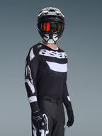 Motorcrosser in de Alpinestars 2026 Racer Air Riway Jersey, zwart-wit shirt met 'astars' logo, bijpassende zwart-witte helm met oranje spiegelende bril, zwarte motorcrossbroek en handschoenen, gecoördineerde outfit voor off-road racen.
