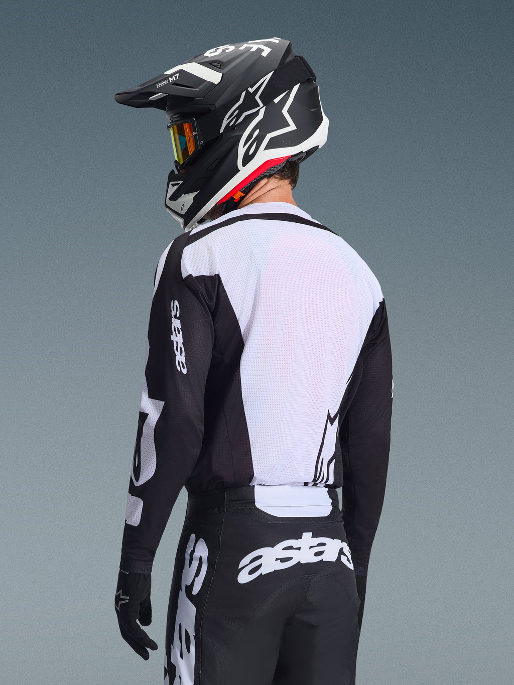 Motocrossrijder van achteren in de Alpinestars 2026 Racer Air Riway Jersey, zwart en wit, met een wit ademend mesh-rugpaneel en zwarte mouwen met 'astars'-branding, bijpassende zwarte broek met witte logo's, zwarte helm met witte accenten, professionele off-road uitrusting voor prestaties bij hoge temperaturen.