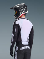 Motocrossrijder van achteren in de Alpinestars 2026 Racer Air Riway Jersey, zwart en wit, met een wit ademend mesh-rugpaneel en zwarte mouwen met 'astars'-branding, bijpassende zwarte broek met witte logo's, zwarte helm met witte accenten, professionele off-road uitrusting voor prestaties bij hoge temperaturen.