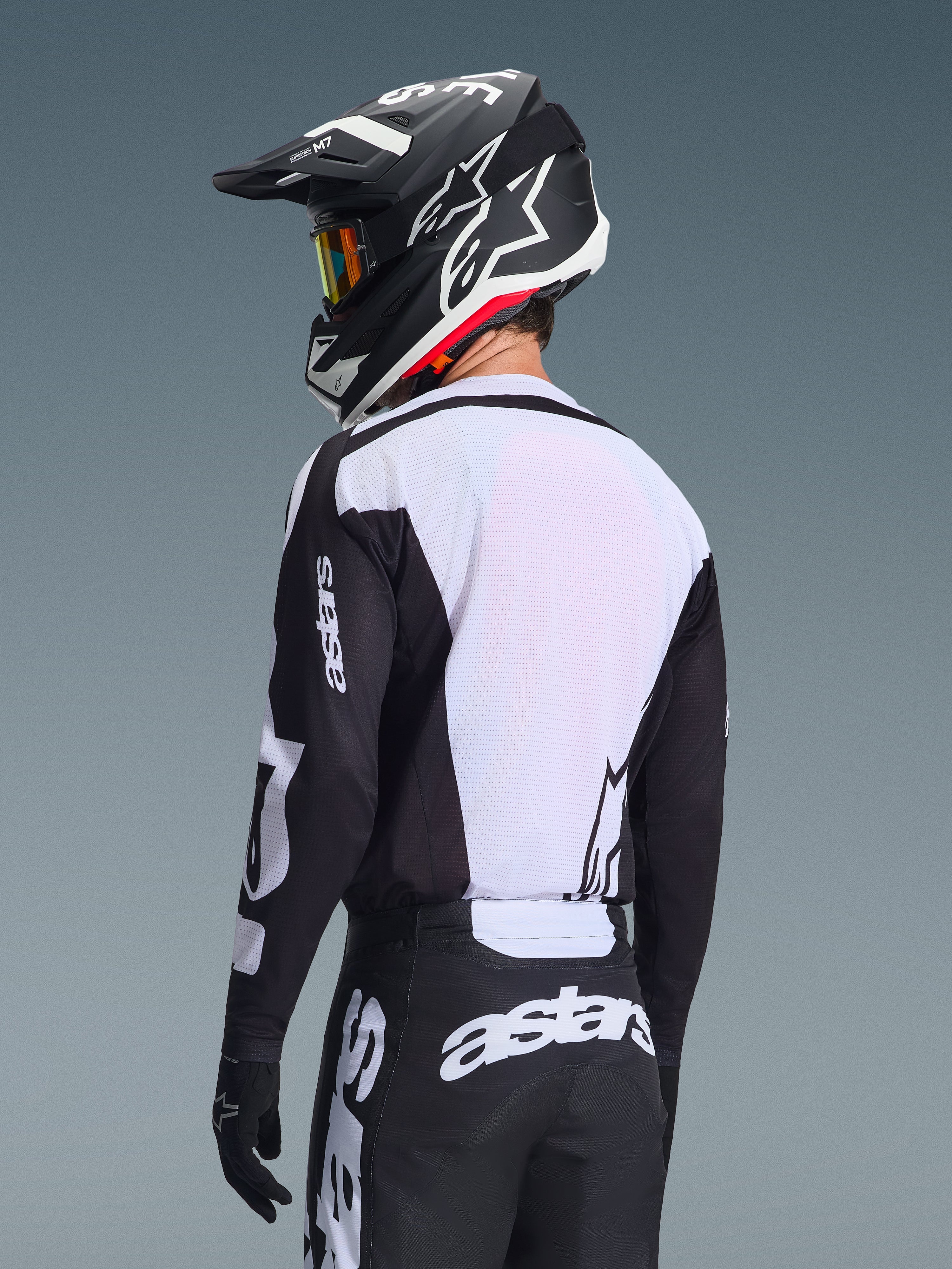 Motocrossrijder van achteren in de Alpinestars 2026 Racer Air Riway Jersey, zwart en wit, met een wit ademend mesh-rugpaneel en zwarte mouwen met 'astars'-branding, bijpassende zwarte broek met witte logo's, zwarte helm met witte accenten, professionele off-road uitrusting voor prestaties bij hoge temperaturen.