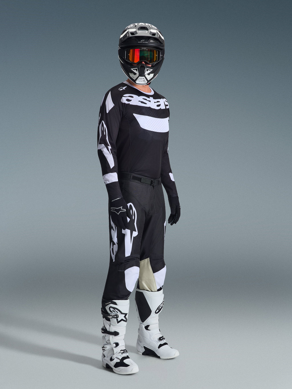 Motorcrossrijder in volledige race-uitrusting, 2026 Racer Air Riway Jersey in zwart en wit, bijpassende zwarte broek met grote witte Alpinestars-logo's, zwarte helm met witte accenten en oranje crossbril, witte en zwarte laarzen, gecoördineerde outfit voor professioneel off-road racen