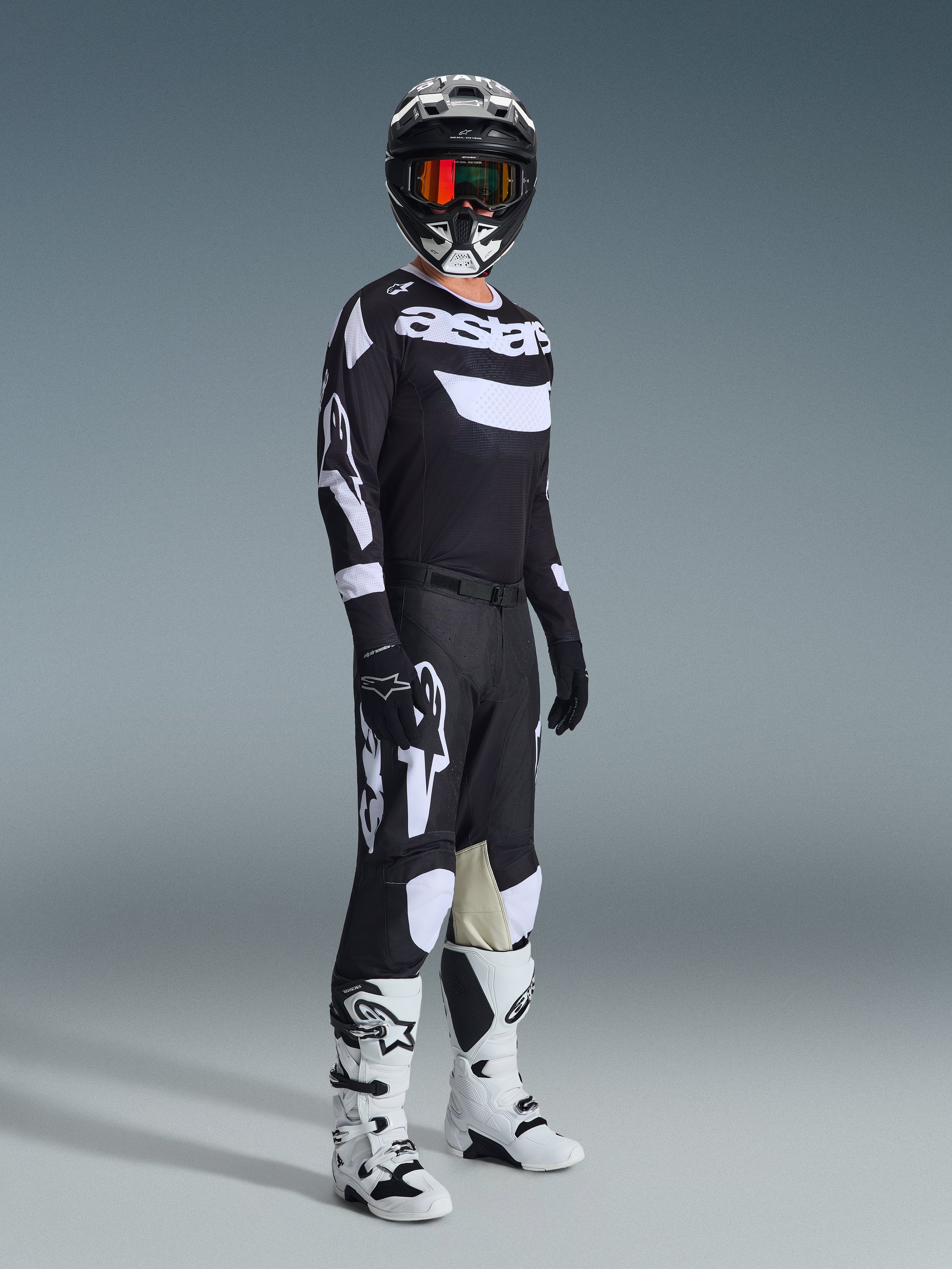 Motorcrossrijder in volledige race-uitrusting, 2026 Racer Air Riway Jersey in zwart en wit, bijpassende zwarte broek met grote witte Alpinestars-logo's, zwarte helm met witte accenten en oranje crossbril, witte en zwarte laarzen, gecoördineerde outfit voor professioneel off-road racen