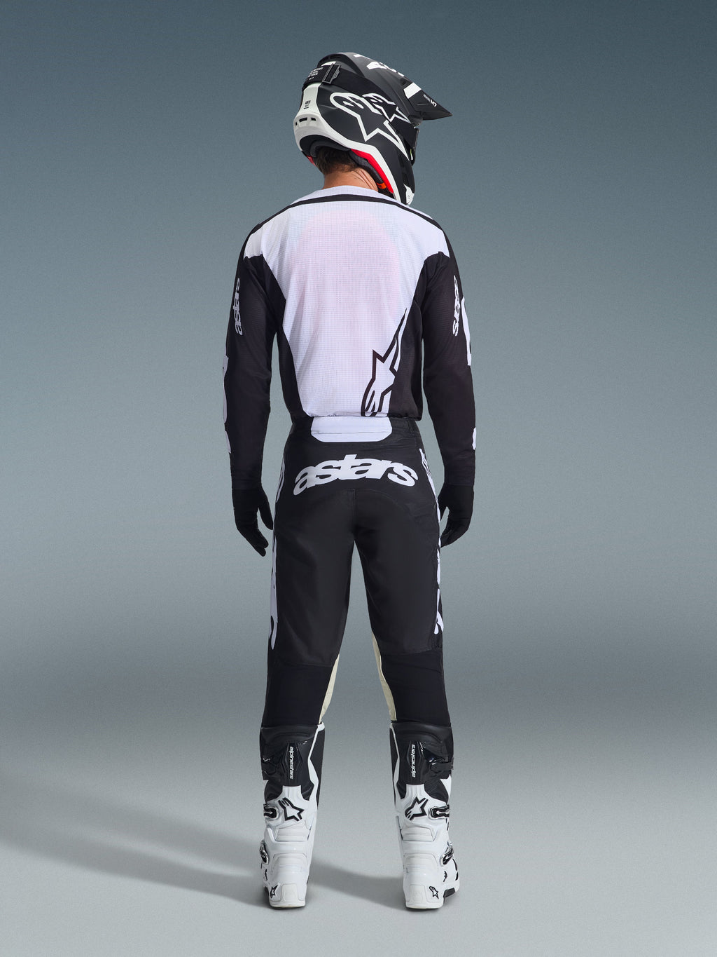 Achteraanzicht van een motorcrosser die de 2026 Racer Air Riway Jersey in zwart en wit draagt, voorzien van een ademend mesh achterpaneel, gecombineerd met een bijpassende zwarte motorcrossbroek met witte 'astars' branding, een zwart-witte helm en witte motorcrosslaarzen.