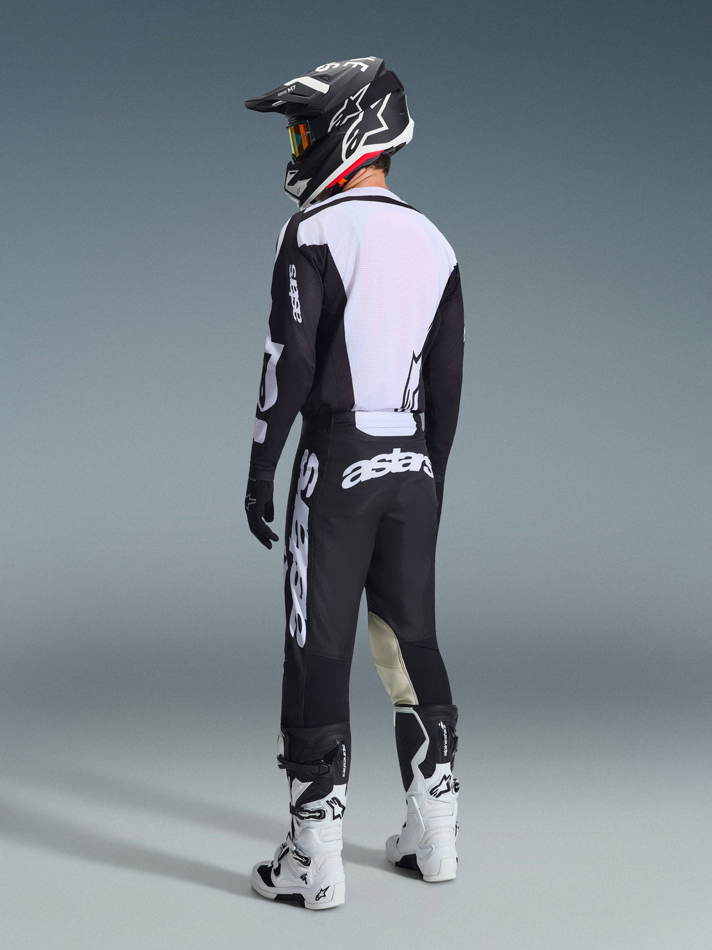 Motorcrosser van achteren gezien in de 2026 Racer Air Riway Jersey in zwart en wit, met het grote witte mesh rugpaneel voor luchtstroom, gecombineerd met een zwarte broek, zwart-witte helm en witte racelaarzen, complete professionele uitrusting voor off-road prestaties.