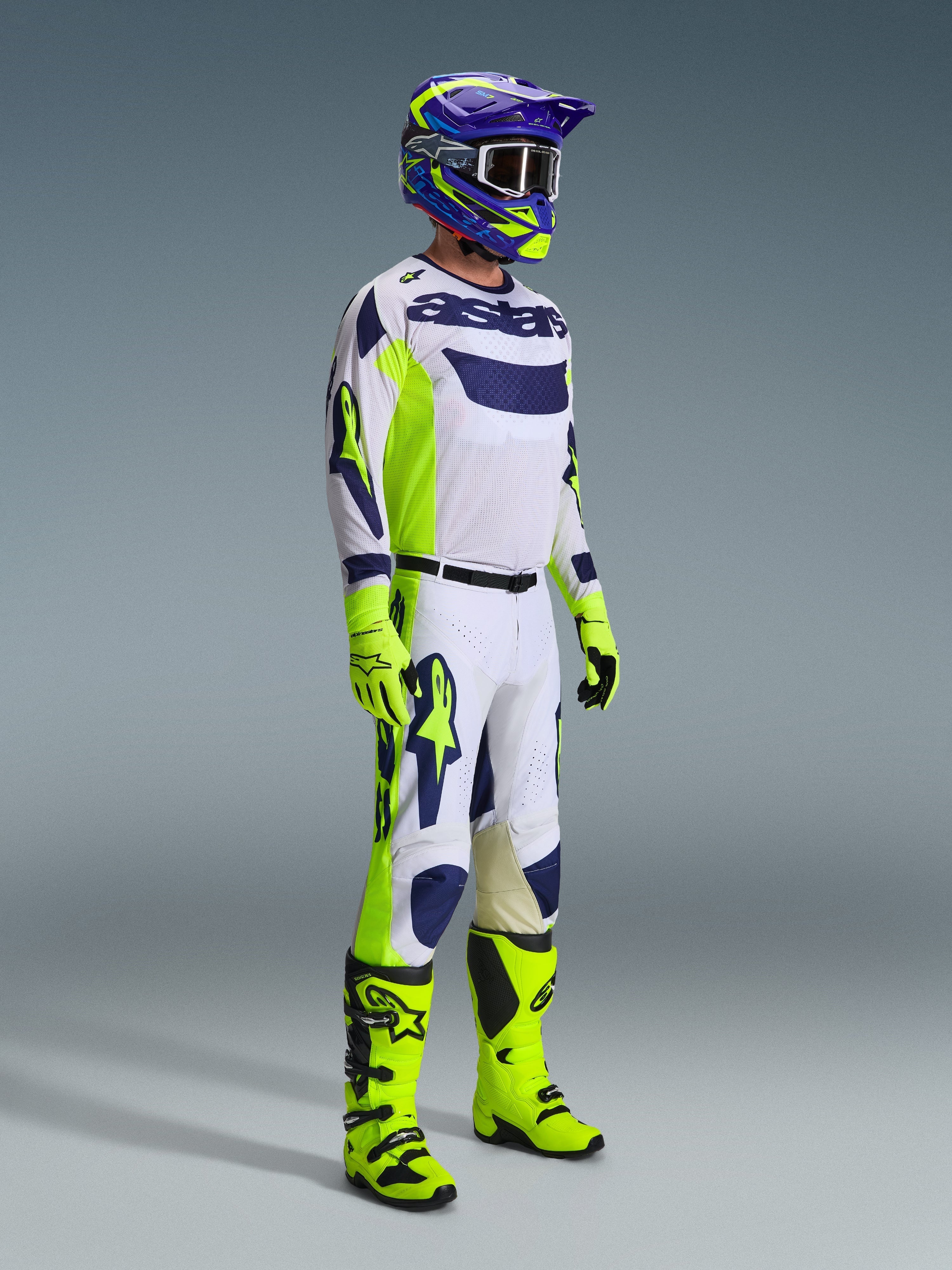 Motocross rider wearing complete racing gear, Light Gray jersey with Yellow Fluo and Blue accents, matching white and yellow pants, bright yellow boots and gloves, purple and yellow helmet with dark goggles, coordinated outfit for off-road racing