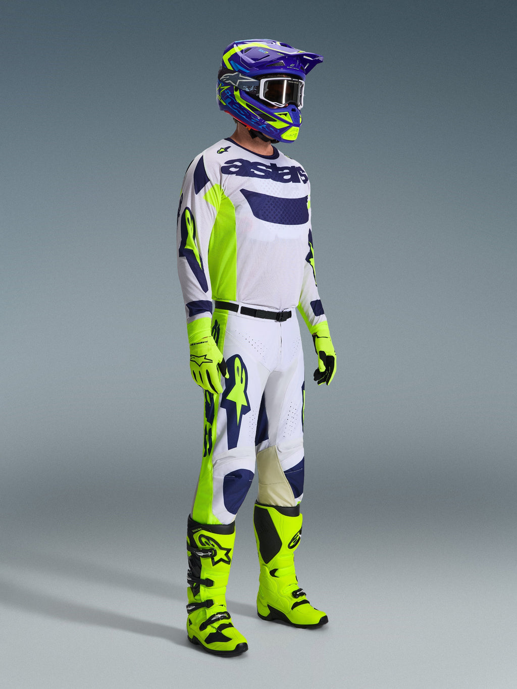 Motocross rider wearing the Alpinestars 2026 Racer Air Riway Gear Set in Light Gray, Yellow Fluo, and Blue. The set includes a long-sleeve mesh jersey, matching racing pants, bright neon yellow gloves and boots, and a purple and blue helmet with goggles, designed for ventilated off-road performance.