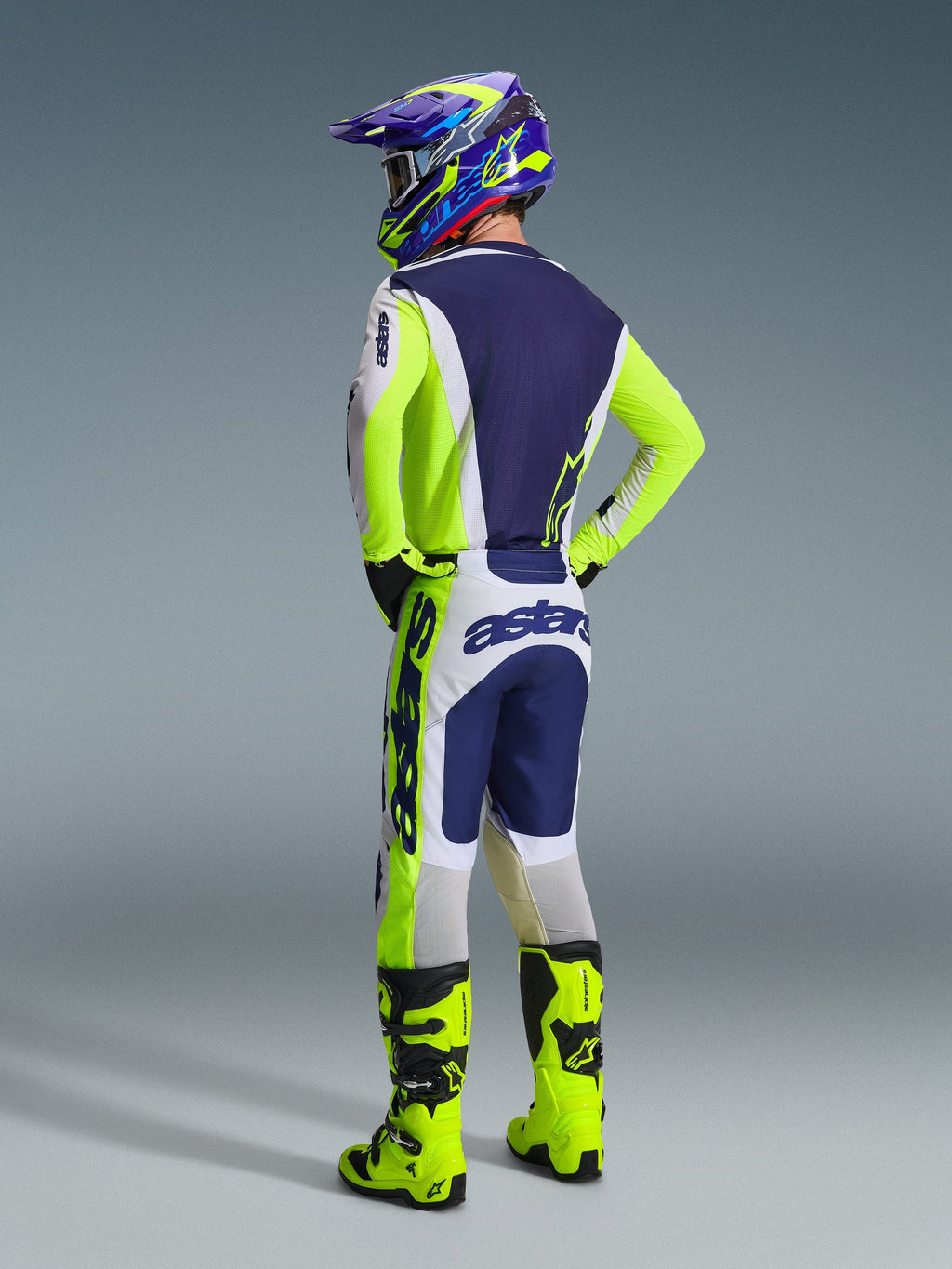 Motocross rider from a back view wearing the Alpinestars 2026 Racer Air Riway Gear Set, Light Gray Yellow Fluo and Blue jersey and pants, featuring 'astars' branding on sleeves and seat, yellow fluo boots, and a matching colorful helmet.