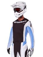 Alpinestars 2025 Techstar Melt Gear Set, motorcrosser in wit met zwarte en lichtblauwe accenten, jersey met zwarte borst en witte mouwen, bijpassende wit-zwarte broek, witte SM10-helm met bril, zwart-witte handschoenen, complete professionele off-road race-outfit.