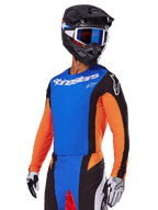 Alpinestars 2025 Techstar Melt Gear Set, motorcrosser met een blauwe jersey met oranje mouwen en zwarte zijpanelen, bijpassende blauwe en zwarte broek, zwarte helm met blauwe bril, witte en zwarte handschoenen, sportieve race-fit motorcrosskleding