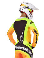Motorcrosser draagt Alpinestars 2025 Techstar Melt Jersey, achteraanzicht, neongeel met oranje mouwen en een groot zwart grafisch paneel, bijpassende crossbroek met opvallende branding, witte Alpinestars helm en oranje crossbril, professionele off-road race-uitrusting