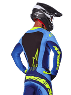 Achteraanzicht van een motorcrossrijder die de Alpinestars 2025 Techstar Melt Jersey draagt in blauw en Yellow Fluo, met een zwart achterpaneel en opvallende gele branding op de mouwen, gecombineerd met een bijpassende crossbroek en zwarte carbon helm, performance off-road racekleding