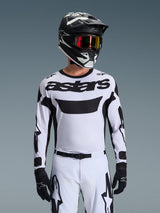 Motocross-rijder in de Alpinestars 2026 Racer Riway Jersey in wit en zwart, voorzien van 'astars'-branding op de borst, gecombineerd met een bijpassende zwart-witte broek, een zwarte helm met bril en zwarte handschoenen, gecoördineerde set voor off-road racen.
