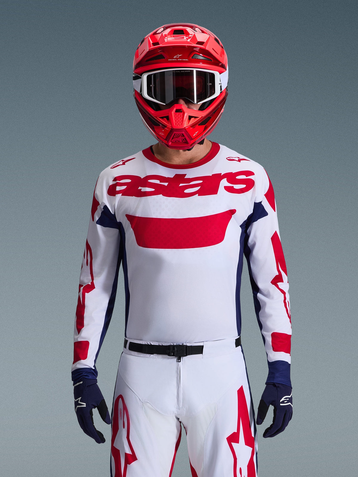 Motocrossrijder met de Alpinestars Racer Riway Jersey 2026 in wit, blauw en rood, voorzien van een groot rood 'astars'-logo op de borst, marineblauwe zijpanelen, bijpassende witte en rode motocrossbroek, een rode helm met witte crossbril en marineblauwe handschoenen, complete off-road race-uitrusting.