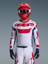 Motocrossrijder met de Alpinestars Racer Riway Jersey 2026 in wit, blauw en rood, voorzien van een groot rood 'astars'-logo op de borst, marineblauwe zijpanelen, bijpassende witte en rode motocrossbroek, een rode helm met witte crossbril en marineblauwe handschoenen, complete off-road race-uitrusting.
