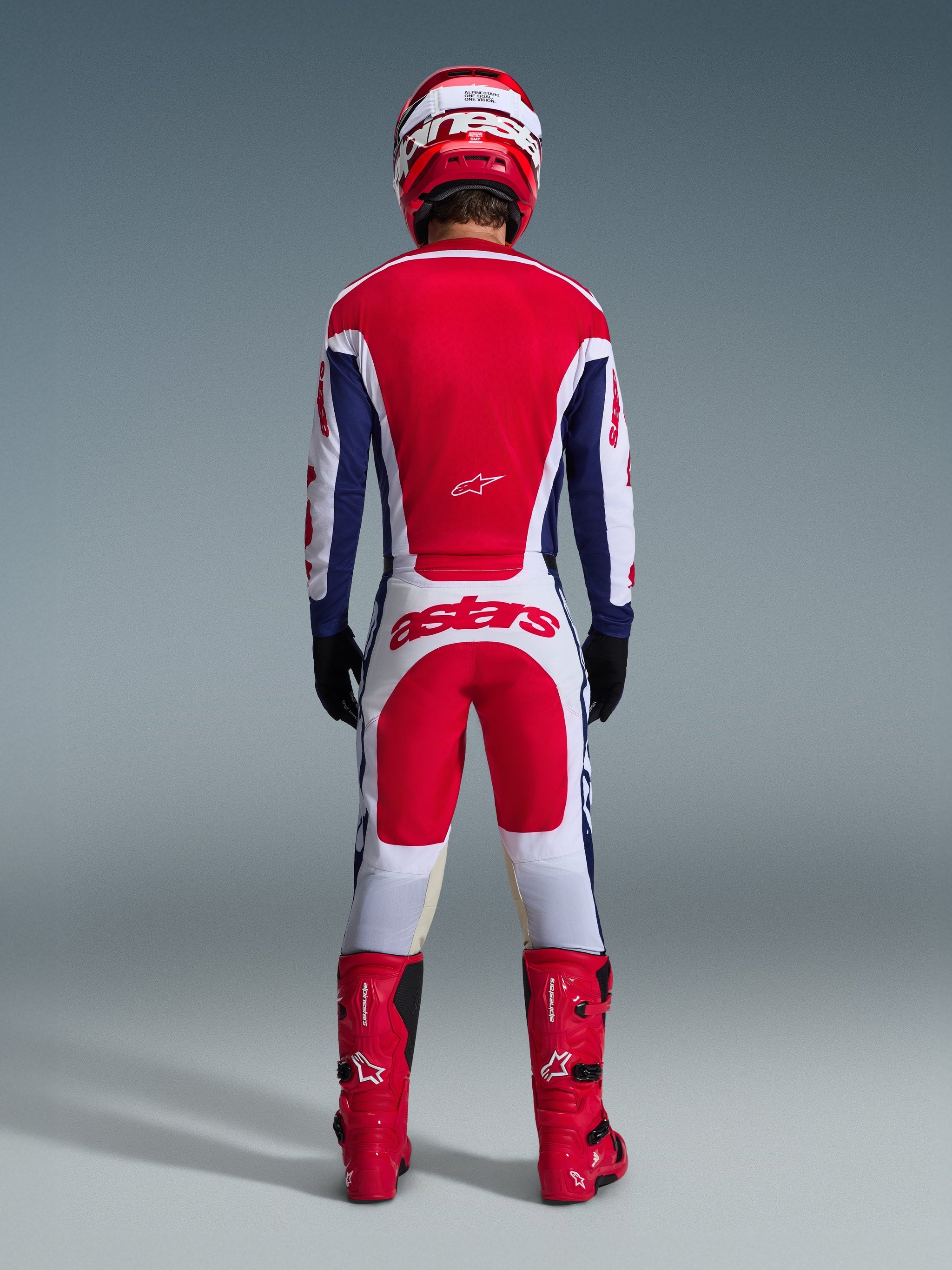 Motorcrossrijder in volledige race-uitrusting van achteren gezien, Alpinestars 2026 Racer Riway Jersey in White Blue Red, bijpassende witte en rode broek met opvallende 'astars' branding, rode laarzen en een rood-witte helm, gecoördineerde off-road race-outfit.