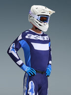 Motorcrosser die de Alpinestars 2026 Racer Riway Jersey in UCLA Blue White draagt, voorzien van 'astars' branding op de borst, gecombineerd met een bijpassende blauwe motorcrossbroek, witte helm met crossbril en blauwe handschoenen, hoogwaardige off-road race-uitrusting