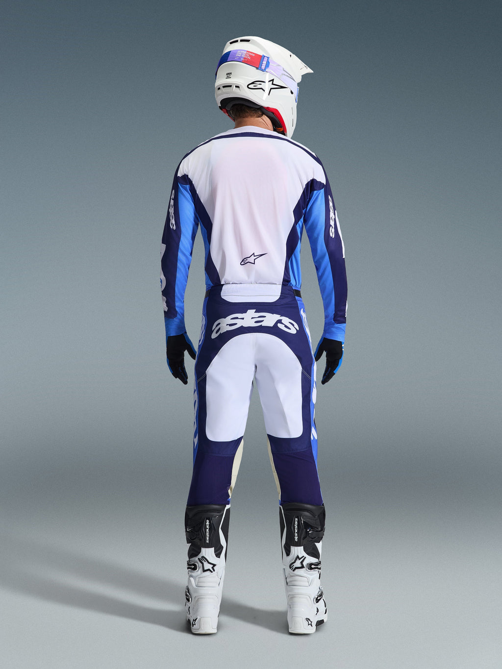 Alpinestars 2026 Racer Riway Jersey in UCLA Blue en wit, motocrosser van achteren gezien in bijpassende broek met witte en marineblauwe panelen en Astars-branding, witte en zwarte laarzen en een witte helm, compleet gecoördineerd racetenue voor off-road prestaties.
