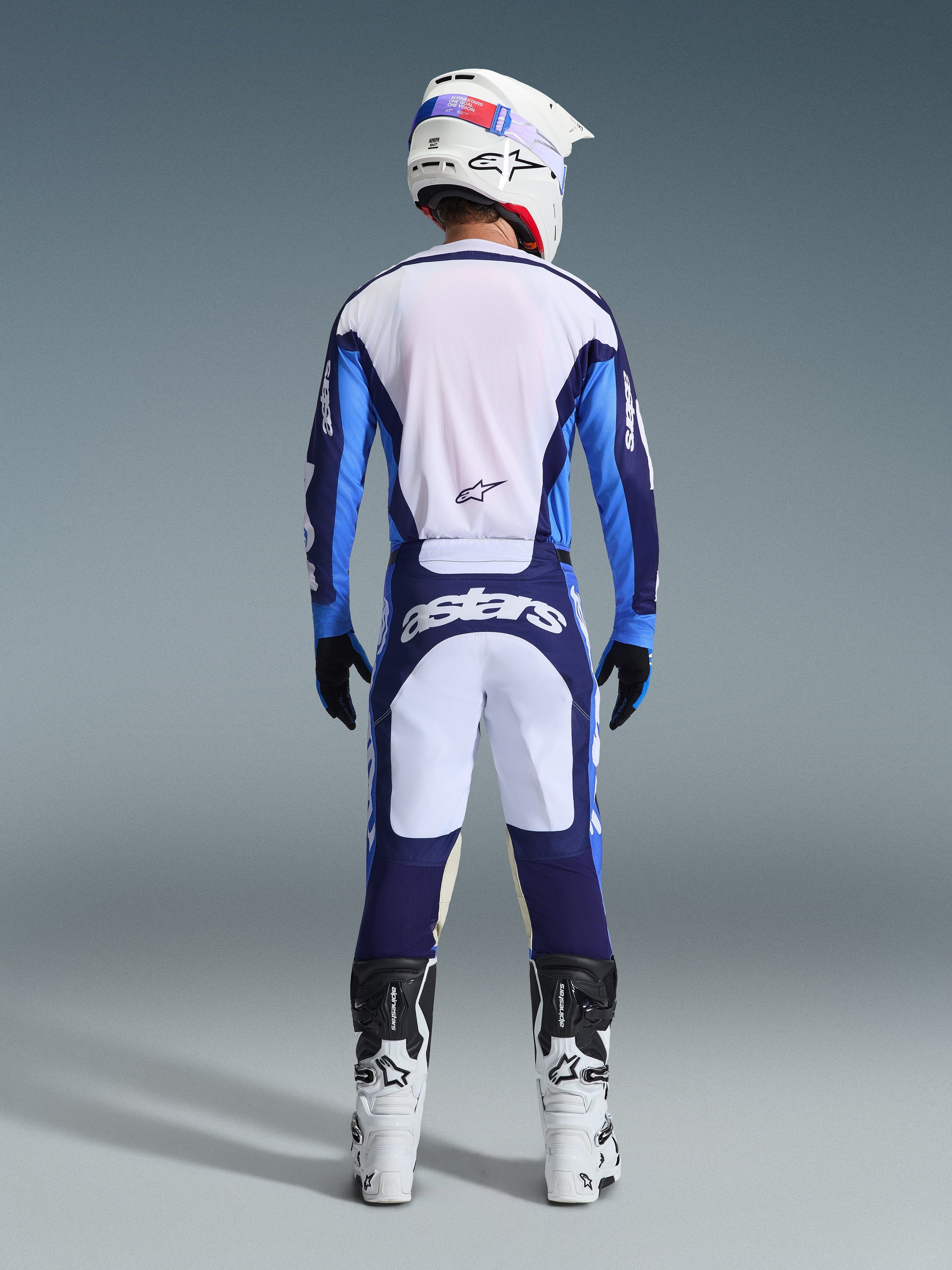 Alpinestars 2026 Racer Riway Jersey in UCLA Blue en wit, motocrosser van achteren gezien in bijpassende broek met witte en marineblauwe panelen en Astars-branding, witte en zwarte laarzen en een witte helm, compleet gecoördineerd racetenue voor off-road prestaties.