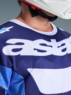 Close-up van Alpinestars 2026 Racer Riway Jersey in Ucla Blue en wit, met een groot wit 'astars'-logo op de borst, witte kraag en ademende mesh-stoftextuur, gedragen met een witte motorcrosshelm