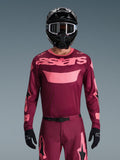 Motorcrosser draagt de Alpinestars 2026 Racer Riway Jersey in Dark Red Coral, met een kastanjebruine body en koraalroze 'astars' logo op de borst, gecombineerd met bijpassende motorcrossbroek, zwarte helm en zwarte handschoenen, professionele uitrusting voor off-road racing.