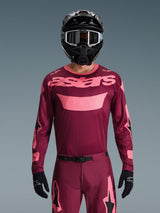 Motorcrosser draagt de Alpinestars 2026 Racer Riway Jersey in Dark Red Coral, met een kastanjebruine body en koraalroze 'astars' logo op de borst, gecombineerd met bijpassende motorcrossbroek, zwarte helm en zwarte handschoenen, professionele uitrusting voor off-road racing.