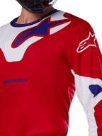 Alpinestars 2025 Racer Veil Jersey, motocross shirt, felrood en wit, close-up van het bovenlichaam en de borst, blauw Alpinestars-logo op een wit kraagpaneel, rode mouwen met witte accenten, gedragen met een zwarte helm, technische off-road kleding