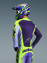 Motocross rider from behind wearing the Alpinestars 2026 Racer Portl Jersey and matching pants, Purple and Yellow Fluo color scheme with lavender accents, coordinated blue and yellow helmet, race-fit gear designed for off-road performance.