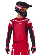 Alpinestars 2025 Fluid Haul Jersey, motorcross shirt, felrood en zwart, gedragen als onderdeel van een volledige set met bijpassende broek en handschoenen, zwarte helm met crossbril met gele lens, witte merklogo's, hoogwaardige off-road racekleding