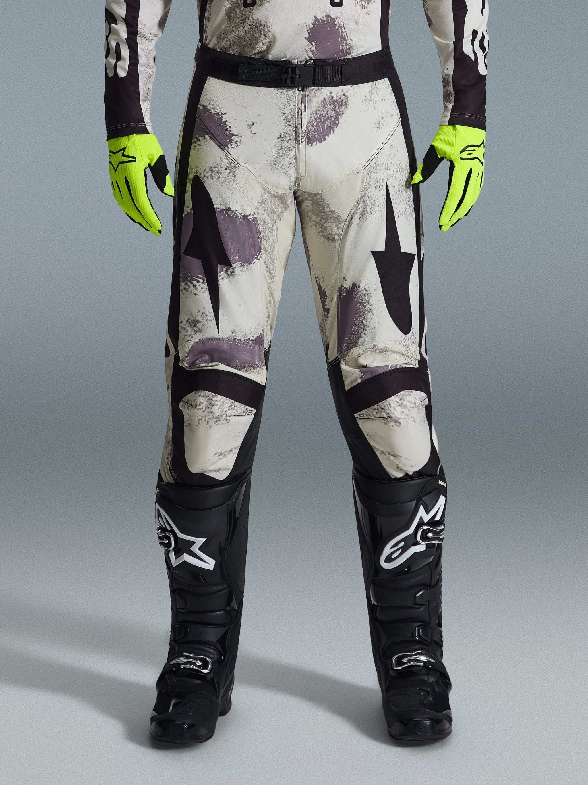 Alpinestars 2026 Racer Lahnd Pants, motorcrossbroek, Sand Camo-patroon met grijze en paarse abstracte graphics, gedragen met neongele handschoenen en zwarte laarzen, voorzien van zwarte sterlogo's op de dijen en een zwarte tailleriem, duurzame off-road race-uitrusting