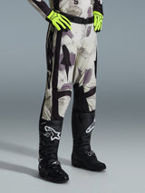 Alpinestars 2026 Racer Lahnd broek, motorcrossbroek, Sand Camo, gedragen met neon gele handschoenen en zwarte motorcrosslaarzen, met een abstract camouflagepatroon en zwarte zijpanelen, ergonomisch off-road ontwerp