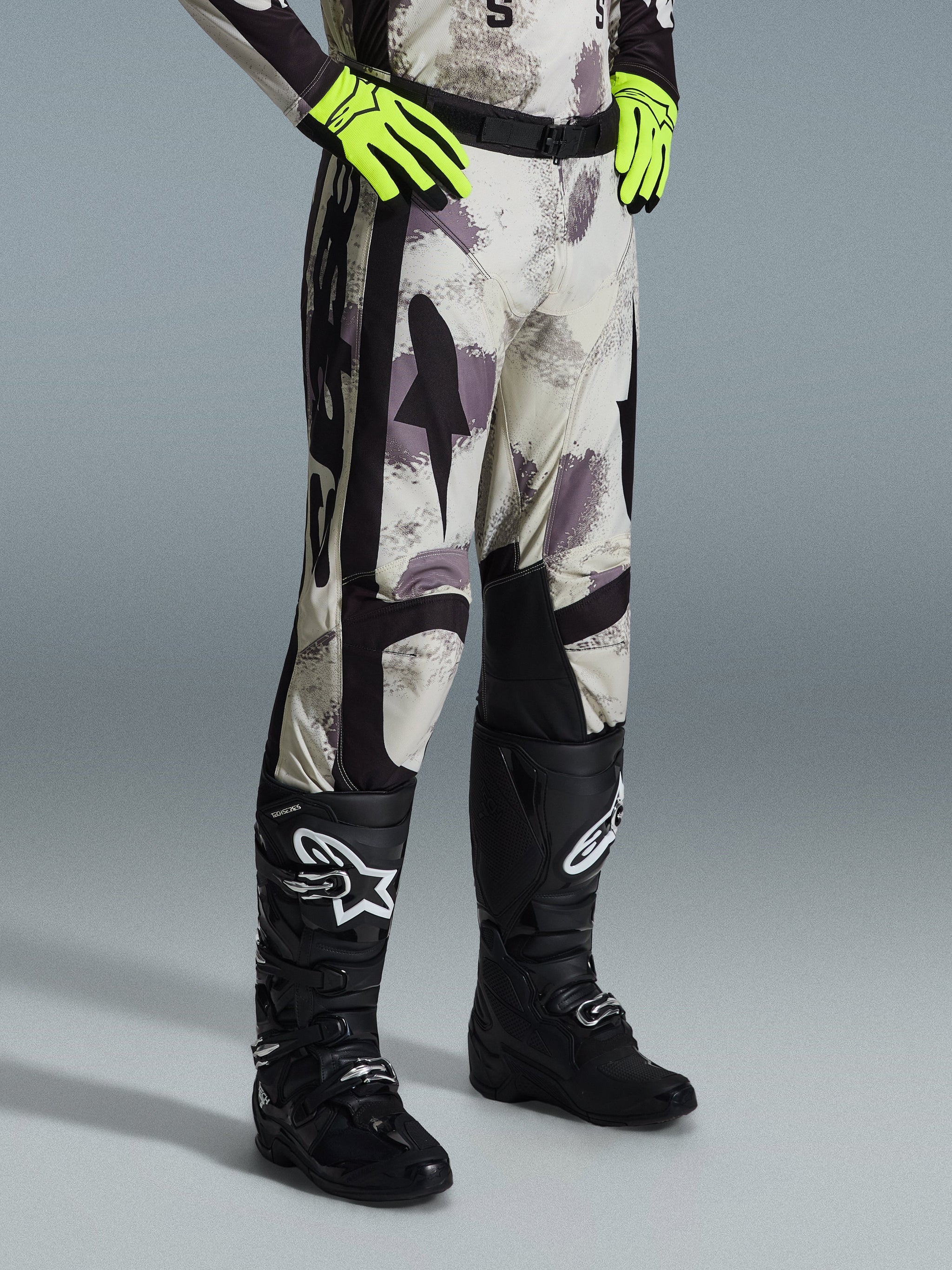 Alpinestars 2026 Racer Lahnd broek, motorcrossbroek, Sand Camo, gedragen met neon gele handschoenen en zwarte motorcrosslaarzen, met een abstract camouflagepatroon en zwarte zijpanelen, ergonomisch off-road ontwerp