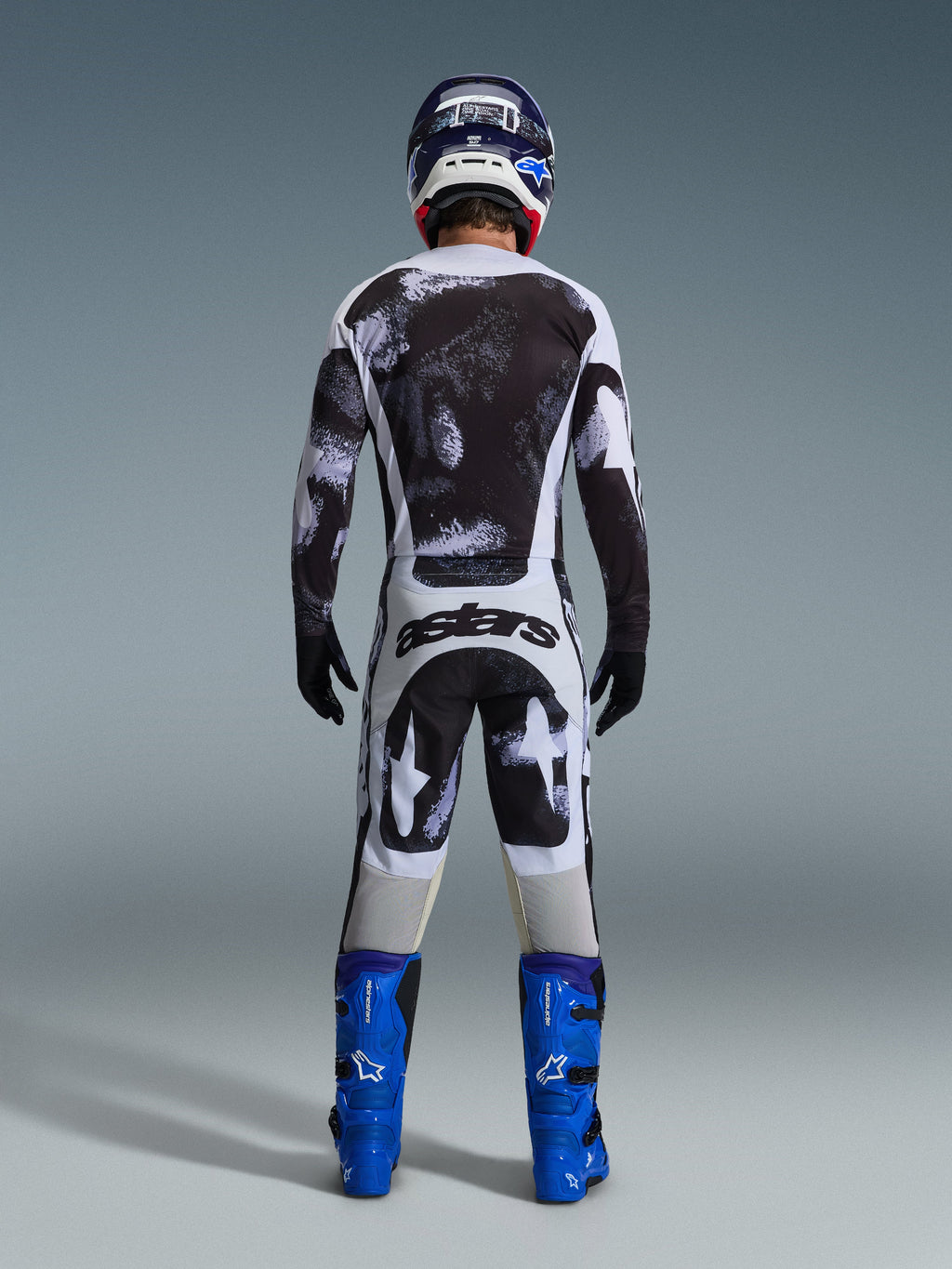 Motocross rider from the rear wearing the Alpinestars 2026 Racer Lahnd Gear Set, black and white abstract patterned jersey and pants, 'astars' branding on the seat, paired with blue boots and a matching helmet, professional off-road racing apparel.