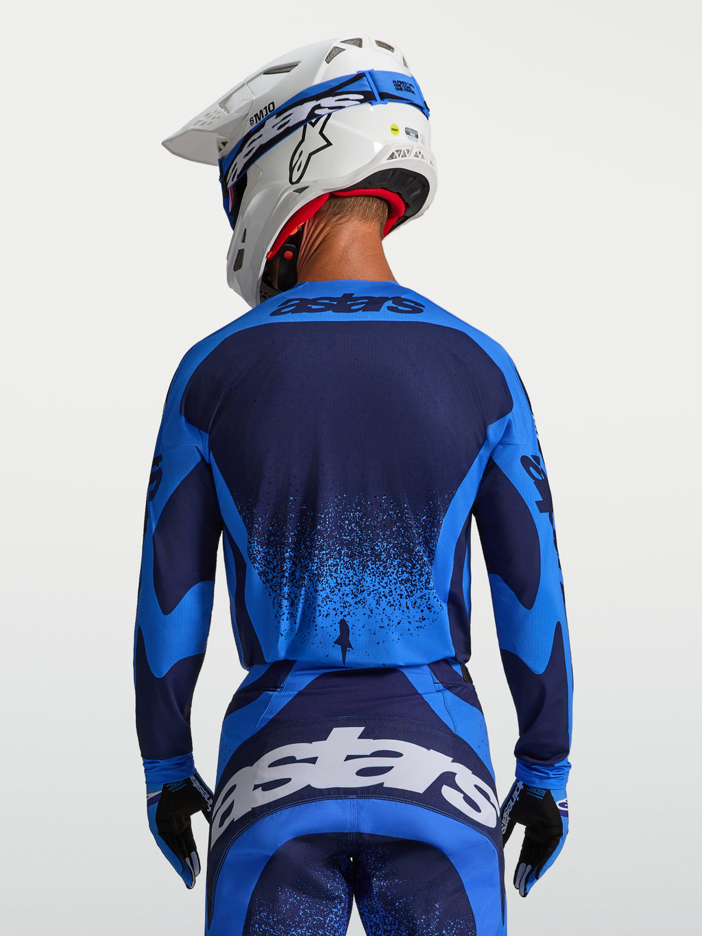 Alpinestars Techstar Swan jersey, heren-motocrossjersey, Ucla blauw en diepblauw, atletische racepasvorm met verlengde achterzoom, achteraanzicht op een model met witte MX-helm.