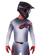 Alpinestars 2025 Supertech Lipan Jersey, motorcross shirt, grijs met oranje en navy blauwe accenten, performance top met lange mouwen en geperforeerde ventilatie, gedragen met zwarte helm en rode handschoenen, strak racing design