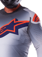 Close-up van een rijder die de Alpinestars 2025 Supertech Lipan Jersey draagt, motorcross jersey, grijs en fluo oranje, voorzien van een groot oranje Alpinestars-logo op de borst, lasergesneden ventilatieopeningen en een zwarte helm, professionele race-uitrusting.