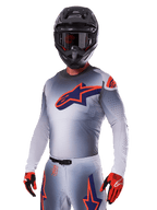 Motorcrosser in Alpinestars 2025 Supertech Lipan Gear Set, grijze jersey en broek met marineblauwe en oranje accenten, matzwarte helm met crossbril en rood-zwarte handschoenen, atletische prestatiekleding voor off-road racen