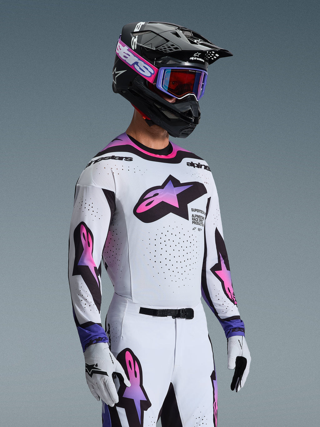 Motocross rider wearing Alpinestars 2026 Supertech AFD Vista Gear Set, white jersey and pants with pink and purple gradient star logos, black helmet with purple accents, purple goggles with red-tinted lenses, and matching white gloves, coordinated professional off-road racing outfit.