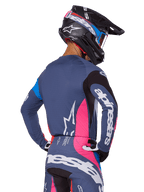 Motorcrosser van achteren gezien in de Alpinestars 2025 Techstar Dreem uitrustingsset in grijs, met een blauwgrijze jersey en broek met roze en zwarte panelen, witte Alpinestars-logo's op de mouwen en rug, en een bijpassende zwarte helm met blauwe en roze graphics.