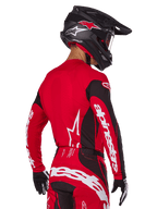 Motorcrosser van achteren gezien in Alpinestars 2025 Techstar Dreem gearset, rood en zwart jersey met witte merknaam op de mouwen, bijpassende rood-zwarte broek met opvallend wit logo op het zitvlak, zwarte carbon helm met rode accenten, zwart-witte handschoenen, professionele off-road outfit.