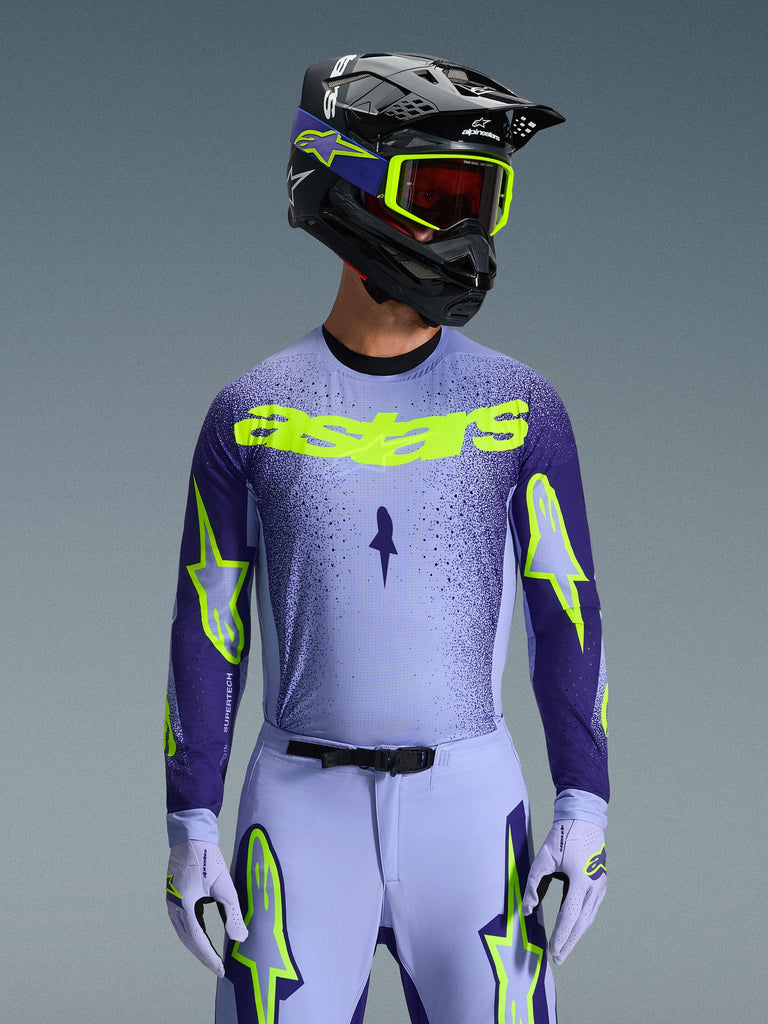Alpinestars 2026 Supertech Scenz Jersey, Motocross Jersey, Purple and Yellow Fluo, worn by a rider with a matching black and purple helmet, neon yellow goggles, and purple racing pants, featuring neon yellow astars branding on the chest and star logos on the sleeves.