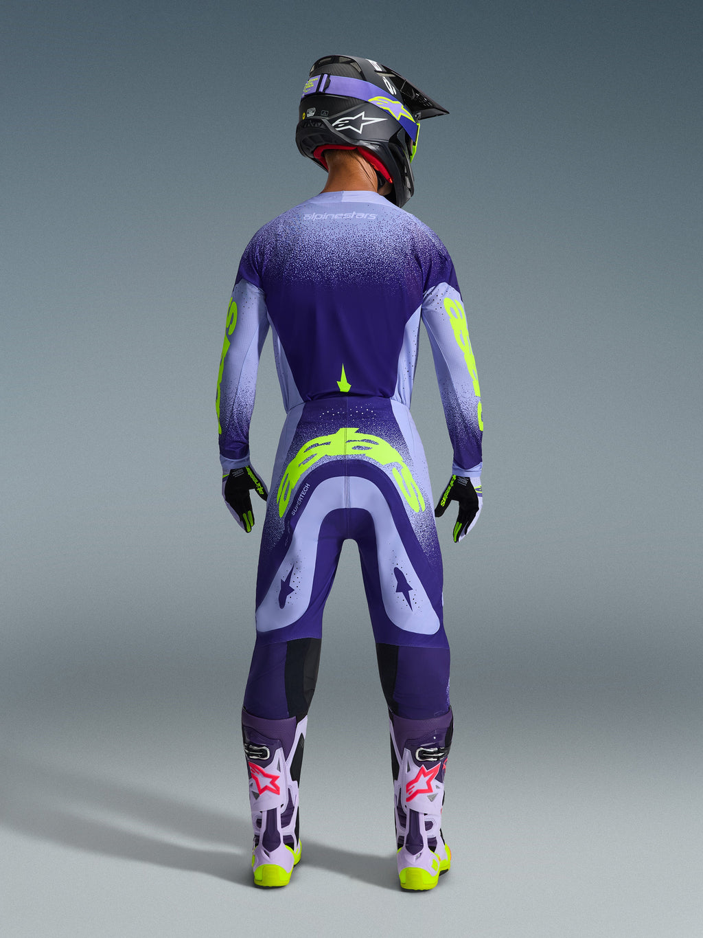 Alpinestars 2026 Supertech Scenz Gear Set, Motocross rider seen from behind wearing purple and lilac gear with neon yellow accents, jersey and pants featuring a gradient dot design, matching helmet and goggles, light purple boots with pink logos and neon yellow soles, professional motocross racing apparel