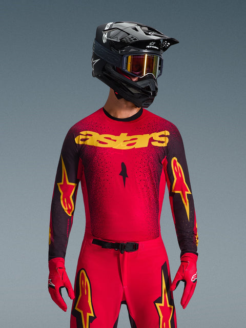 Alpinestars 2026 Supertech Scenz Jersey, motorcross racing shirt, rood en zwart, met gele astars-tekst en sterrenlogo's, lange mouwen, gladde glanzende textuur, gedragen met zwarte helm en rode goggle