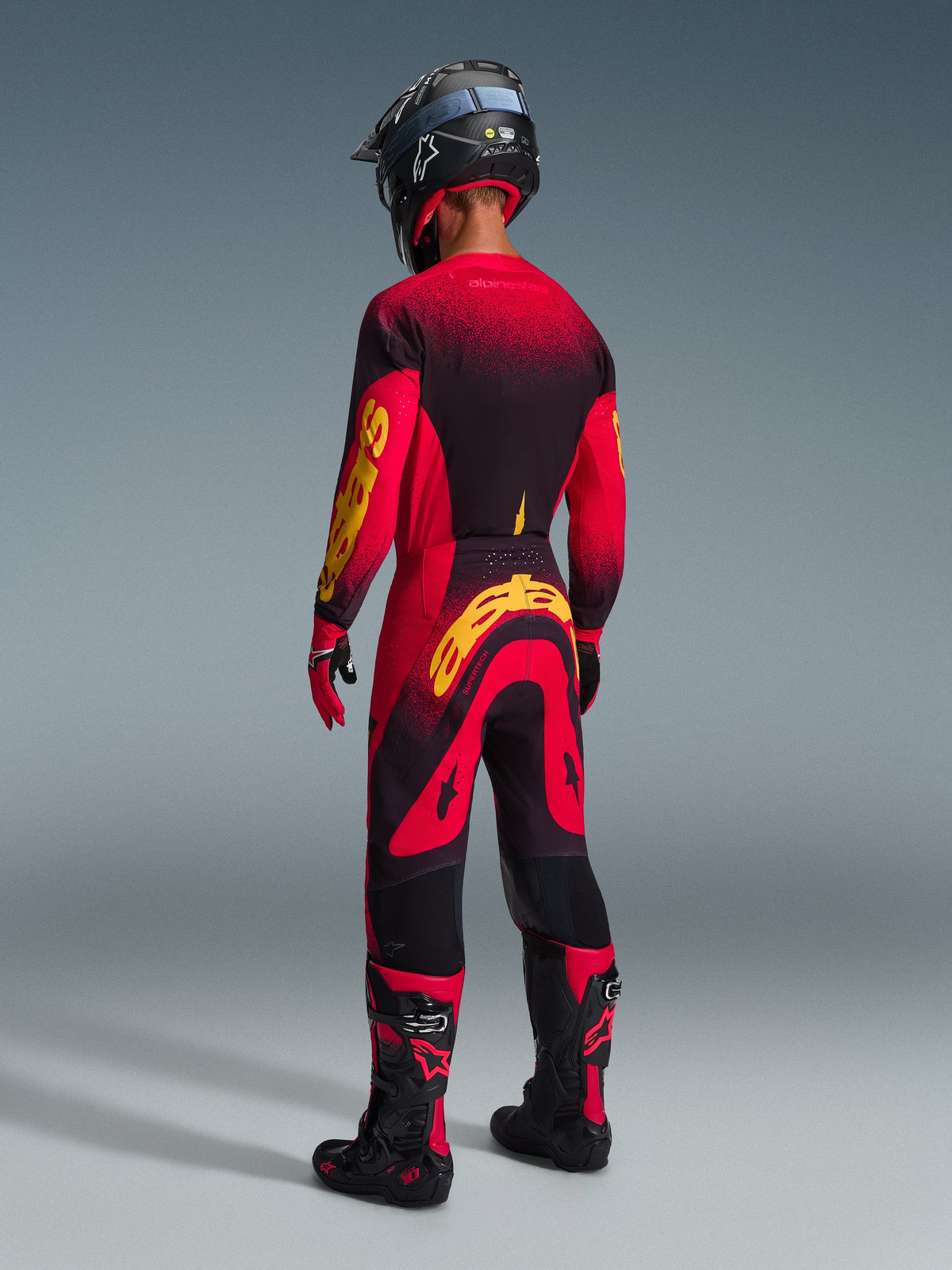 Motorcrosser van achteren gezien in volledige race-uitrusting, rood jersey met zwarte en gele accenten, bijpassende rood-zwarte broek met groot geel Alpinestars-logo, zwarte helm, zwart-rode laarzen en rood-zwarte handschoenen, gecoördineerde outfit voor professionele off-road racing