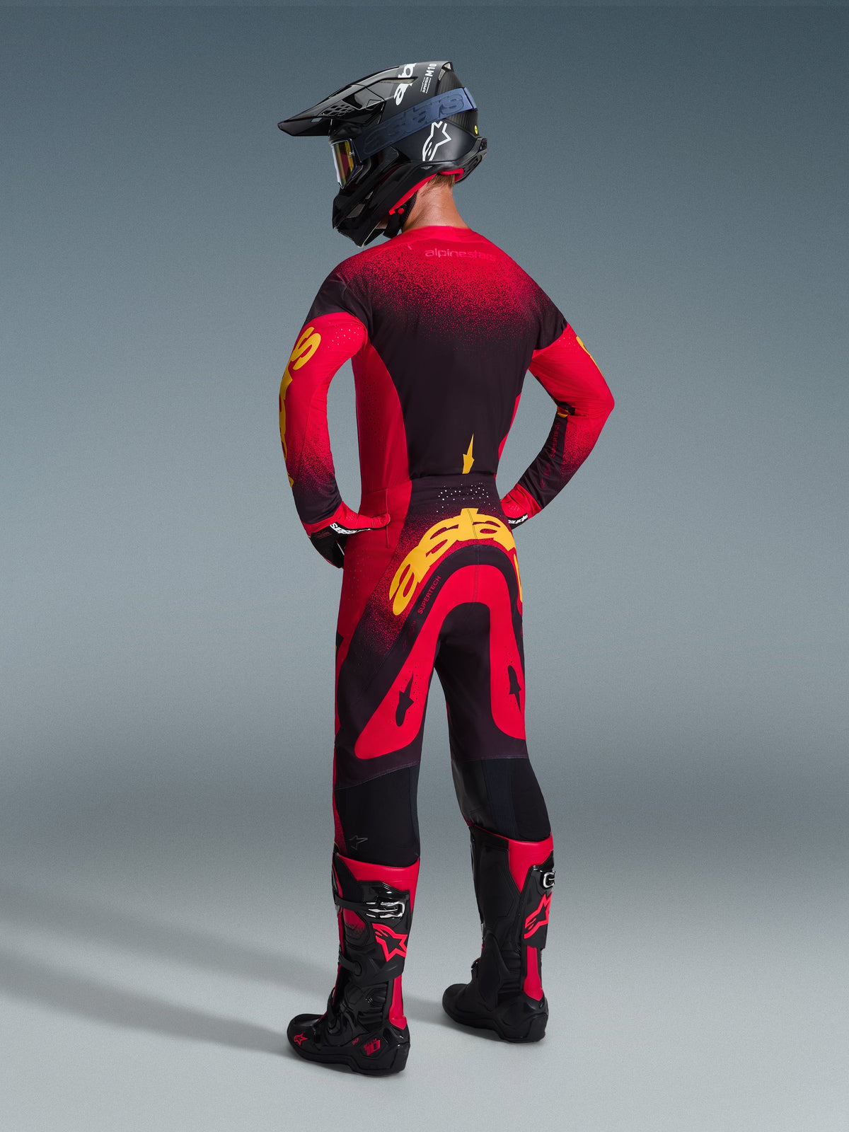 Motocrossrijder met volledige race-uitrusting van achteren gezien, Alpinestars 2026 Supertech Scenz Jersey in rood, zwart en geel, bijpassende motorcrossbroek met gele branding, zwarte helm en zwart-rode laarzen, gecoördineerde outfit voor professioneel off-road racen.