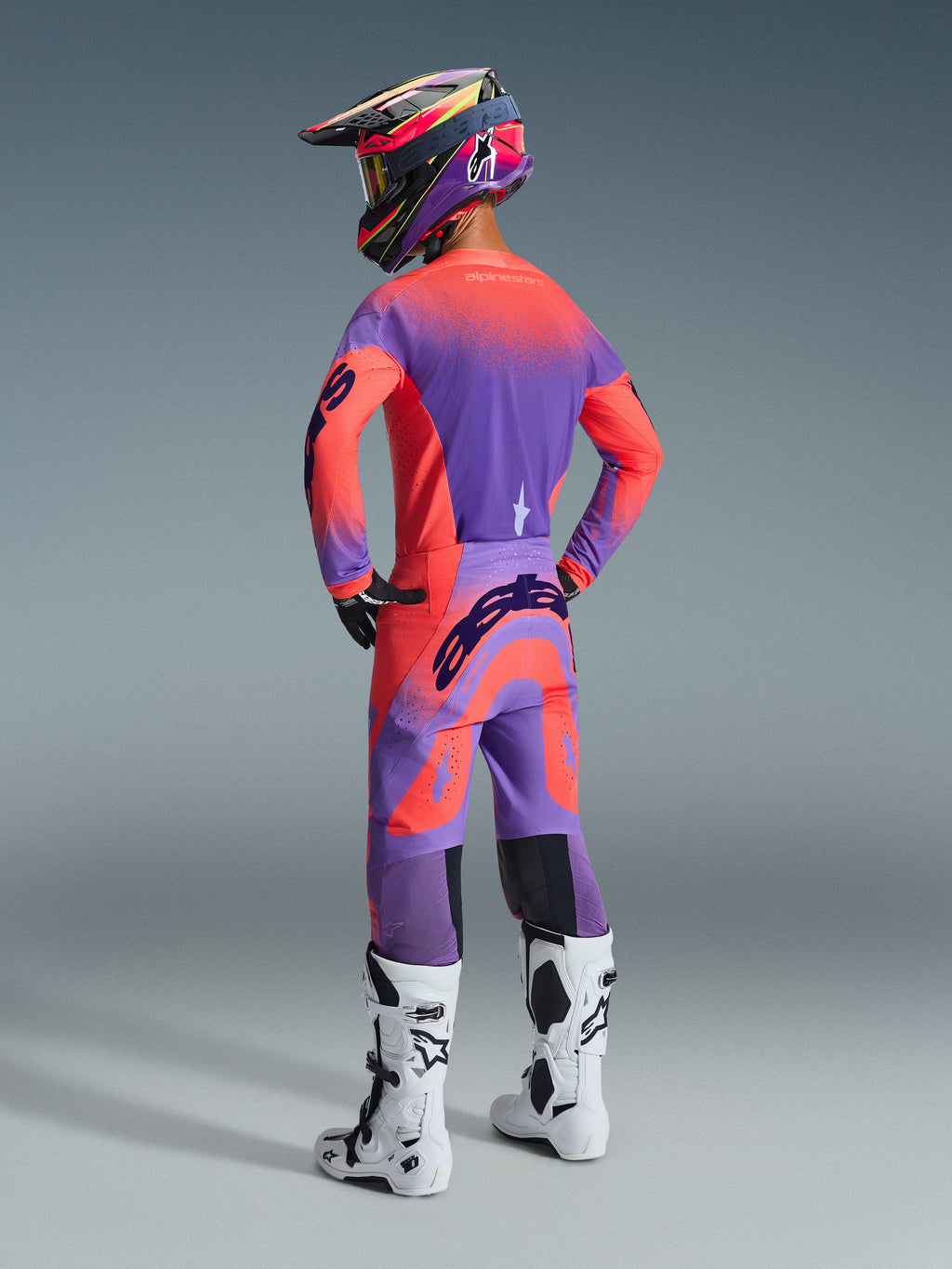 Motocross rider seen from the back wearing the Alpinestars 2026 Supertech Scenz Gear Set, purple and coral pink jersey and pants, large Alpinestars branding on the seat and sleeves, matching purple and coral helmet, white and black boots, coordinated professional racing outfit